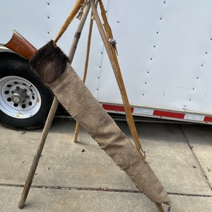 May include: A brown leather quiver with a fur flap, hanging from a wooden tripod stand. The quiver is decorated with beads and has a fringe at the bottom.