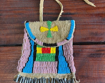 May include: A beaded leather pouch with a fringe. The pouch is decorated with a cross design in yellow, green, and blue beads. The pouch is made of brown leather and has a light brown strap.