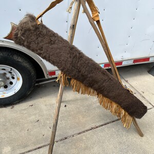 May include: A brown leather gun case with a fringe trim and a strap. The case is made of buffalo hide and is displayed on a wooden tripod.
