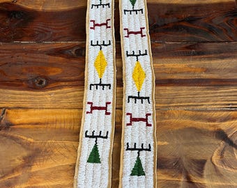 May include: Two beaded strips of leather with geometric designs in white, green, yellow, red, and black beads. The strips are on a wooden surface.