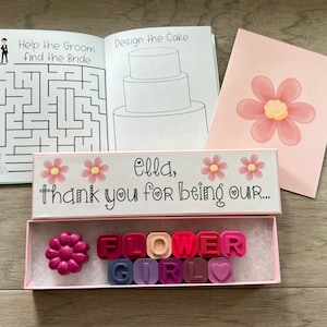 May include: A pink box with a white lid that says "Ella, thank you for being our..." with two pink flowers on the lid. The box contains a set of colorful flower girl crayons in a pink and white box. The crayons spell out "FLOWER GIRL" with a heart.