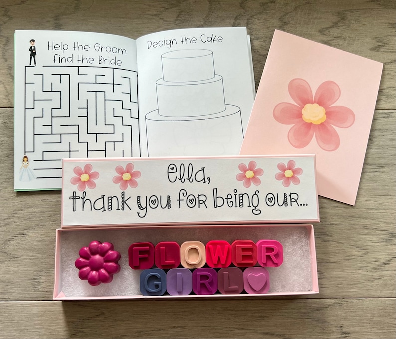 May include: A pink box with a white lid that says "Ella, thank you for being our..." with two pink flowers on the lid. The box contains a set of colorful flower girl crayons in a pink and white box. The crayons spell out "FLOWER GIRL" with a heart.