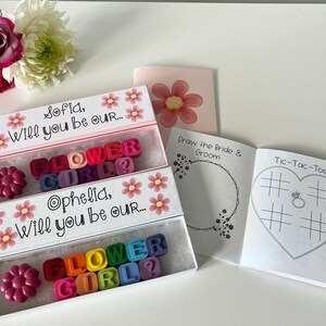 May include: Two white boxes with pink lids and floral designs. Each box contains a message asking "Will you be our... Flower Girl?" with colorful letter blocks. A small white booklet with a heart-shaped tic-tac-toe game and a drawing page for the bride and groom.
