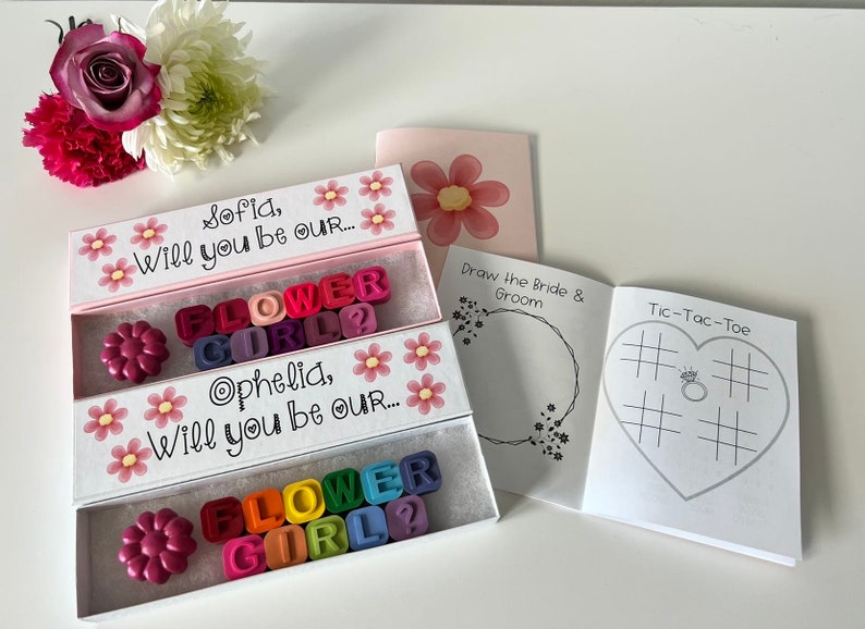 May include: Two white boxes with pink lids and floral designs. Each box contains a message asking "Will you be our... Flower Girl?" with colorful letter blocks. A small white booklet with a heart-shaped tic-tac-toe game and a drawing page for the bride and groom.