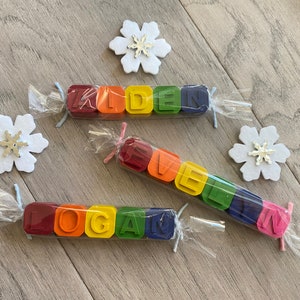 May include: Three sets of colourful crayon blocks spelling out the names "Kiden", "Logan", and "Evelyn". Each set is wrapped in clear plastic and tied with a ribbon.