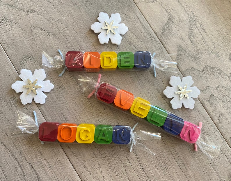 May include: Three sets of colourful crayon blocks spelling out the names "Kiden", "Logan", and "Evelyn". Each set is wrapped in clear plastic and tied with a ribbon.