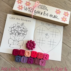 May include: A craft kit for a flower girl with a brown box, a small notebook, crayons, and a tic-tac-toe game. The box has pink flowers and the text "ellie, will you be our..." on it.