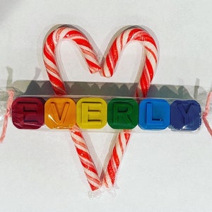 May include: A set of colourful alphabet blocks spelling the name "EVERLY" is wrapped in clear cellophane with a pink ribbon. Two candy canes are tied together in a heart shape behind the blocks.