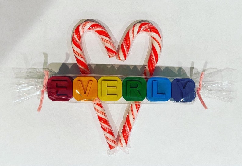 May include: A set of colourful alphabet blocks spelling the name "EVERLY" is wrapped in clear cellophane with a pink ribbon. Two candy canes are tied together in a heart shape behind the blocks.
