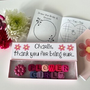 May include: A pink box with the text "Charlie, thank you for being our... FLOWER GIRL" and a flower-shaped crayon inside. The box is surrounded by flowers and a small booklet with the text "Draw the Bride & Groom" and "Tic-Tac-Toe".