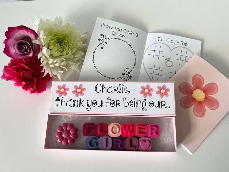 May include: A pink box with the text "Charlie, thank you for being our... FLOWER GIRL" and a flower-shaped crayon inside. The box is surrounded by flowers and a small booklet with the text "Draw the Bride & Groom" and "Tic-Tac-Toe".