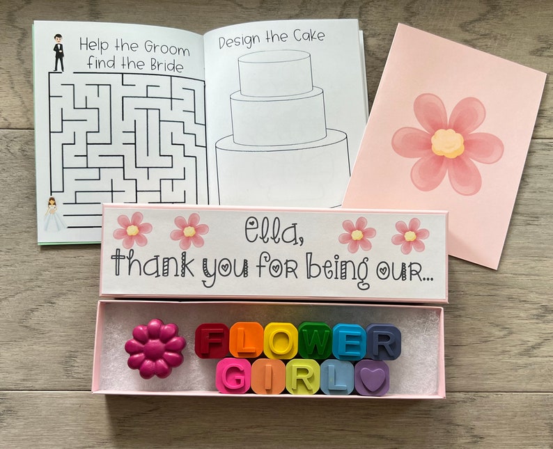 May include: A pink box with the text "Ella, thank you for being our..." and two pink flowers. The box contains a pink flower-shaped crayon and a set of rainbow crayons that spell out "FLOWER GIRL".