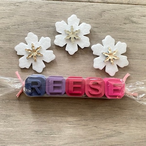 May include: A set of five colourful wax crayons shaped like blocks, spelling out the name "REESE". The crayons are wrapped in clear plastic with a pink ribbon. Three white felt snowflakes with gold glitter centres are in the background.