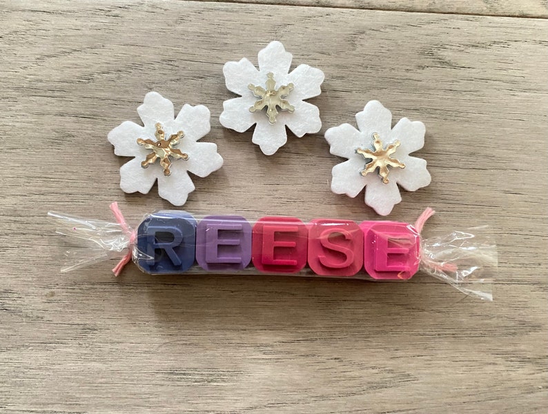 May include: A set of five colourful wax crayons shaped like blocks, spelling out the name "REESE". The crayons are wrapped in clear plastic with a pink ribbon. Three white felt snowflakes with gold glitter centres are in the background.