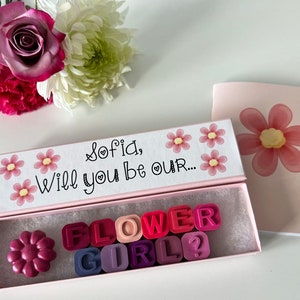 May include: A pink box with a white lid that says "Sofia, Will you be our..." with pink flowers. The box is open and contains pink, red, and purple letter tiles that spell out "FLOWER GIRL?" and a single pink flower.