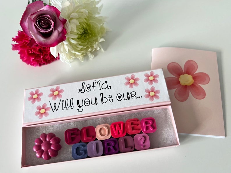 May include: A pink box with a white lid that says "Sofia, Will you be our..." with pink flowers. The box is open and contains pink, red, and purple letter tiles that spell out "FLOWER GIRL?" and a single pink flower.