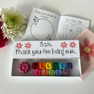 May include: A white box with a pink flower design and the text "Jade, thank you for being our..."  The box contains a set of colorful letter crayons that spell out "FLOWER GIRL".