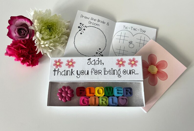 May include: A white box with a pink flower design and the text "Jade, thank you for being our..."  The box contains a set of colorful letter crayons that spell out "FLOWER GIRL".