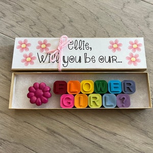 May include: A small wooden box with a pink and white floral design. The box is open and contains a flower-shaped crayon and a set of rainbow-colored crayons that spell out "FLOWER GIRL?"