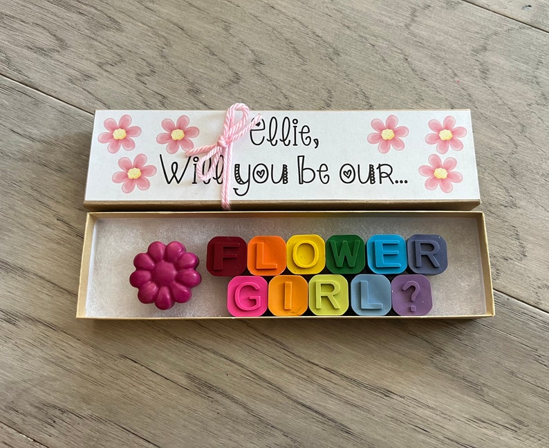 May include: A small wooden box with a pink and white floral design. The box is open and contains a flower-shaped crayon and a set of rainbow-colored crayons that spell out "FLOWER GIRL?"