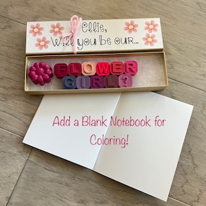 May include: A white box with pink flowers and the text "Ellie, Will you be our... FLOWER GIRL?"  The box contains colorful wax letters spelling out the words "FLOWER GIRL".