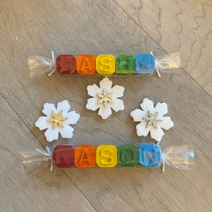May include: Two sets of colourful crayon blocks spelling out the names "CASH" and "KASON" are wrapped in clear plastic with white string. Three white felt snowflakes with silver glitter centres are on a wood background.