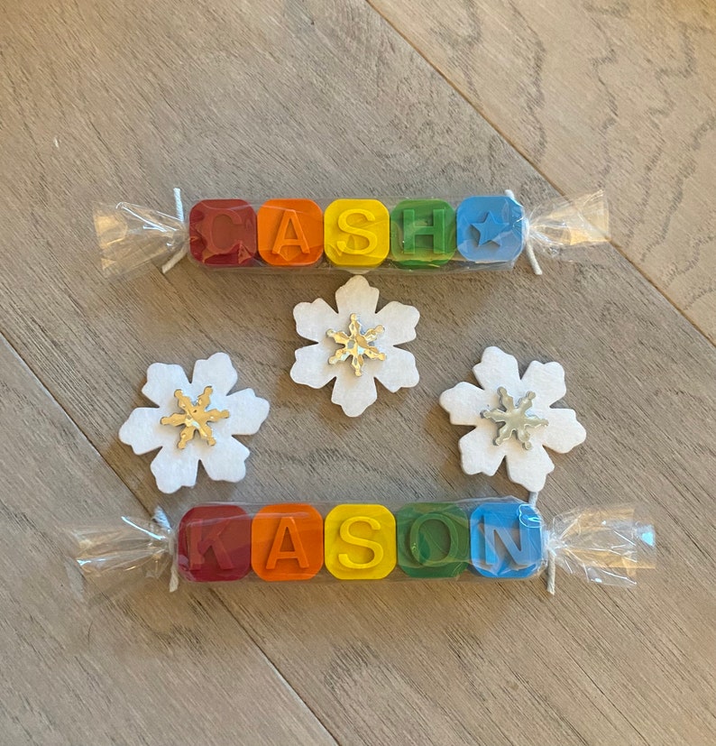 May include: Two sets of colourful crayon blocks spelling out the names "CASH" and "KASON" are wrapped in clear plastic with white string. Three white felt snowflakes with silver glitter centres are on a wood background.