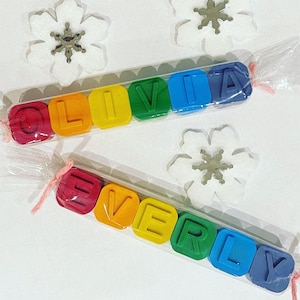 May include: Two sets of colourful crayon blocks spelling out the names "Olivia" and "Everly". The blocks are wrapped in clear plastic and tied with pink ribbon.