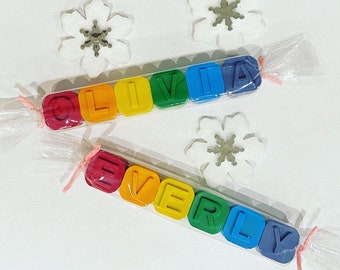 May include: Two sets of colorful crayon blocks spelling out the names "Olivia" and "Everly". The blocks are wrapped in clear plastic and tied with pink ribbon.