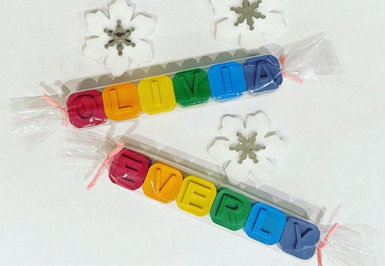 May include: Two sets of colourful crayon blocks spelling out the names "Olivia" and "Everly". The blocks are wrapped in clear plastic and tied with pink ribbon.