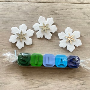 May include: A set of six colourful crayons shaped like the letters S, I, L, A, and S. The crayons are wrapped in clear plastic and tied with a white string. Three white felt snowflakes with silver glitter snowflakes in the centre are on a wooden surface.