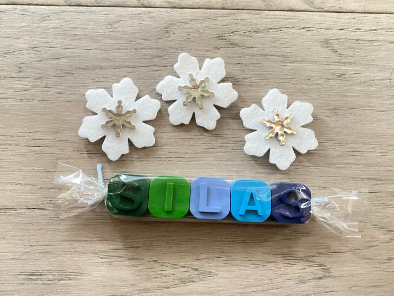May include: A set of six colourful crayons shaped like the letters S, I, L, A, and S. The crayons are wrapped in clear plastic and tied with a white string. Three white felt snowflakes with silver glitter snowflakes in the centre are on a wooden surface.