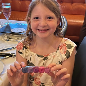 May include: A young person wearing a white dress with pink and green floral print holds a clear plastic bag with colourful letter blocks spelling "VEDA".