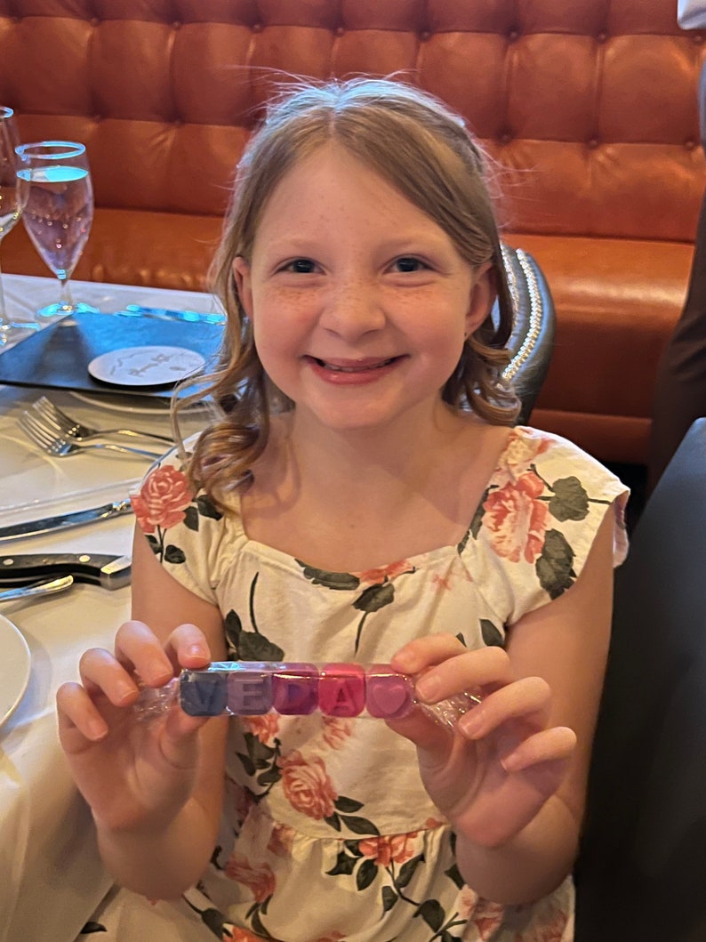 May include: A young person wearing a white dress with pink and green floral print holds a clear plastic bag with colourful letter blocks spelling "VEDA".