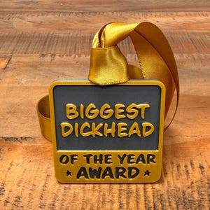 May include: A gold and black award plaque with the text "Biggest Dickhead of the Year Award" on a wooden surface.