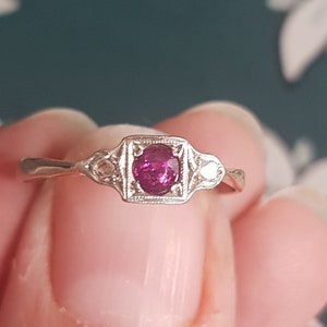 May include: A silver ring with a central, square-set, oval-cut ruby gemstone. The ring has a delicate band and is flanked by two smaller, clear gemstones. The ring is held between fingers against a floral background.