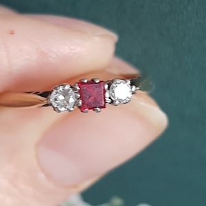 Puede incluir: Un anillo de oro con una gema roja cuadrada flanqueada por dos gemas redondas y transparentes. El anillo se sostiene sobre un fondo verde oscuro con un estampado floral. Una joya clásica.