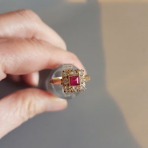 May include: A gold ring featuring a square red gemstone surrounded by smaller clear gemstones.