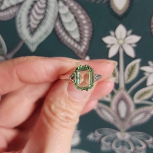 Op de afbeelding: Een zilveren ring met een grote, rechthoekige, lichtgroene edelsteen. De ring heeft ingewikkelde filigraan details op de band en rond de steen. De edelsteen is facetgeslepen en reflecteert licht. De ring wordt vastgehouden.