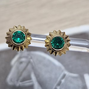 May include: A pair of stud earrings featuring round, vibrant green gemstones set in gold-toned metal. Each earring has a distinctive sunburst or star-shaped border with pointed rays surrounding the central stone, creating a textured, metallic frame.