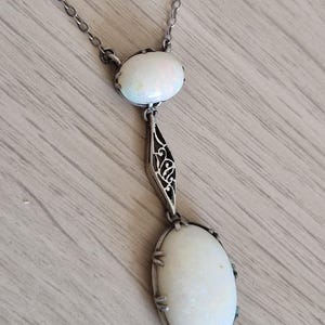 Könnte beinhalten: Eine silberfarbene Halskette mit zwei ovalen, weißen Opalen in Metallfassungen. Der obere, kleinere Opal ist durch ein zentrales, silberfarbenes Metallglied mit einem filigranen, durchbrochenen Muster mit dem unteren, größeren Opal verbunden.