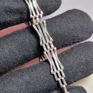 Puede incluir: Una pulsera de plata con un diseño único. La pulsera presenta una serie de barras paralelas interconectadas que crean un efecto tejido. El metal tiene un acabado brillante y reflectante. La pulsera se muestra sobre un fondo oscuro.