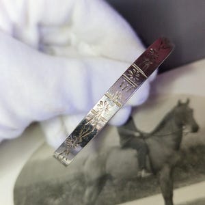 以下が含まれることがあります： 花柄のシルバーカラーのバングルブレスレット。ブレスレットは長方形で、表面にはエッチングされた花の模様があります。背景には、馬とライダーの白黒写真があります。