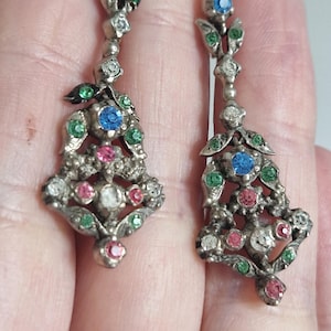 May include: A pair of silver-toned drop earrings featuring an intricate metalwork design adorned with small, multi-colored rhinestones in shades of blue, green, pink, and clear. The earrings have a dangling, elongated shape with floral motifs.