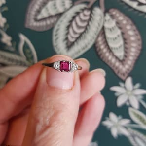 Pode incluir: Um anel de prata com uma pedra vermelha quadrada e duas pedras brancas menores de cada lado.