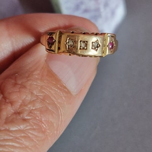 May include: A gold ring with a series of square and star-shaped settings. The ring features two red gemstones and three clear gemstones. The ring is held between fingers.