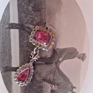 Op de afbeelding: Een vintage hanger met twee roze edelstenen. De bovenste steen is rechthoekig en de onderste druppelvormig, beide omgeven door kleinere, amberkleurige stenen. De hanger hangt aan een zilverkleurige ketting met decoratieve versieringen.
