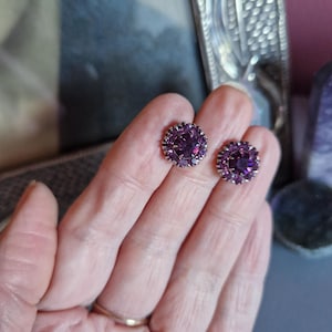 Może przedstawiać: Para kolczyków z fioletowymi kryształami i srebrnymi oprawami. Kolczyki są okrągłe i mają skupisko kryształów w środku.