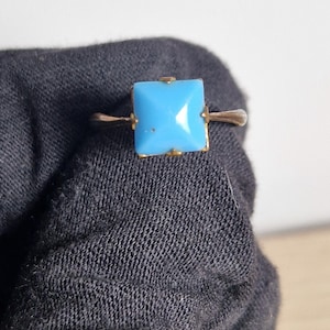 May include: A gold ring with a square, light blue gemstone. The gemstone is set in a gold band with delicate detailing. The ring is a classic piece of jewellery.
