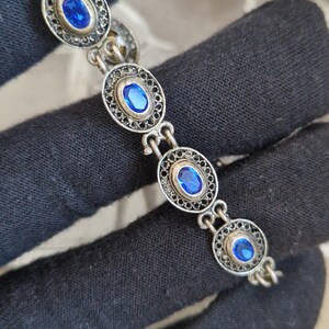 May include: A silver-toned link bracelet featuring a series of oval-shaped settings. Each setting contains a vibrant, oval-cut blue stone surrounded by a delicate, intricate filigree metal border. The links are connected by small silver rings.